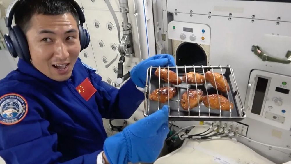 Un astronaute chinois souriant montre un plateau d'ailes de poulet cuites au four dans une station spatiale, portant des écouteurs et des gants bleus.