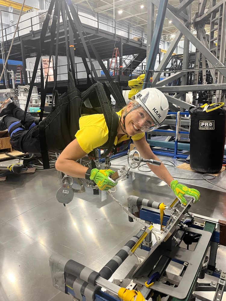 Sophie Adenot, en harnais suspendu, portant un casque et des gants, travaille sur une structure métallique dans un environnement industriel.