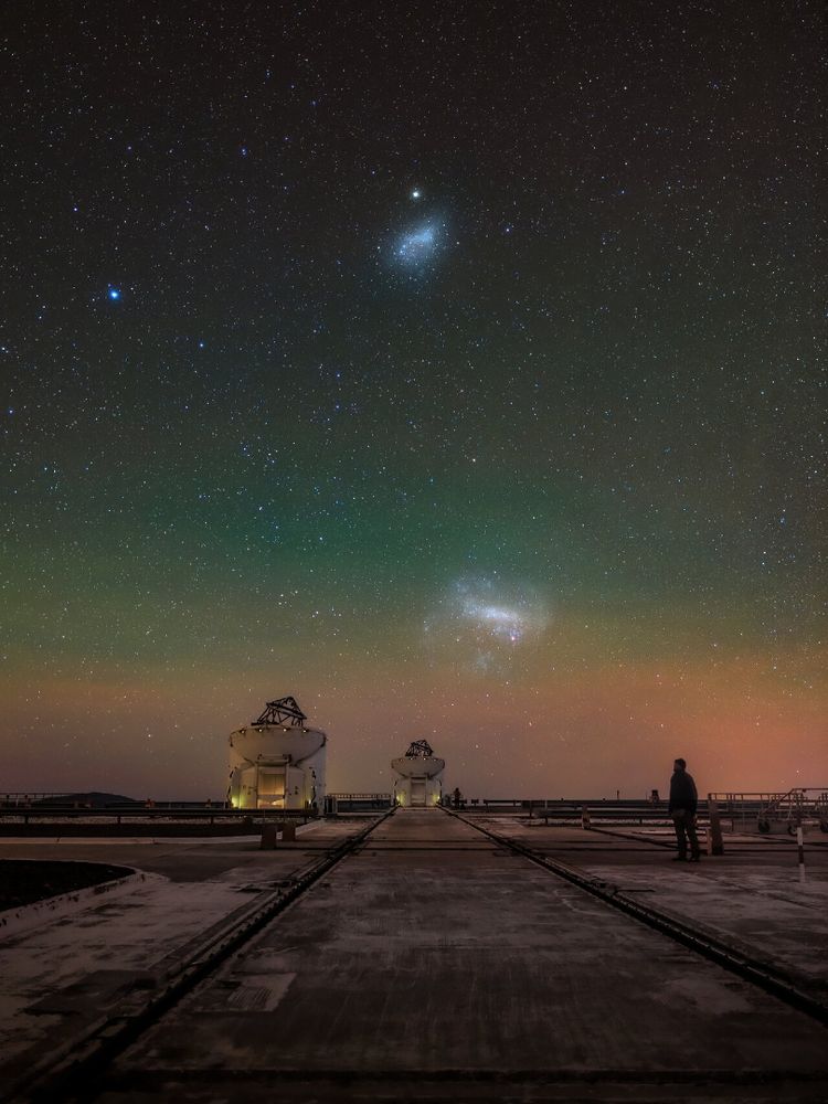 Un ciel étoilé avec deux galaxies visibles au-dessus d'un observatoire. Une personne se tient à droite, regardant les étoiles.