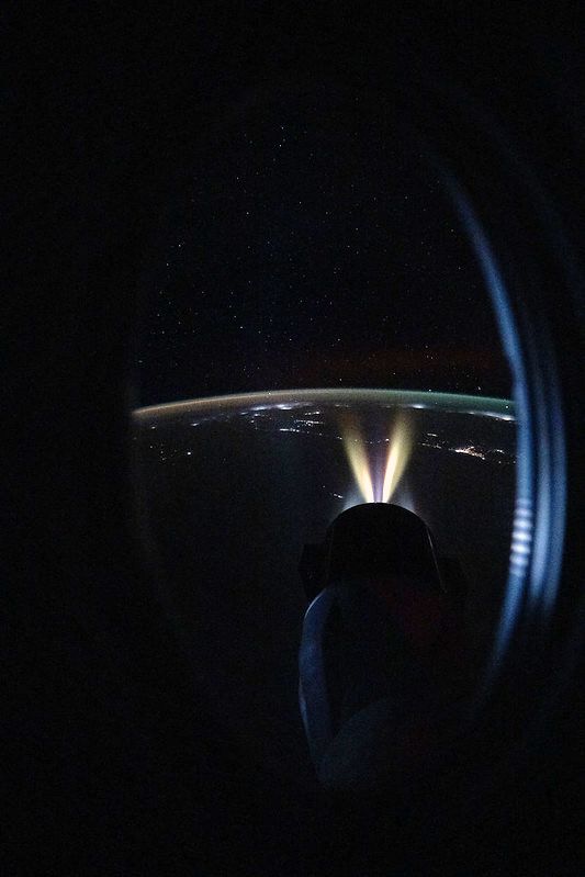 Vue depuis un hublot du Crew Dragon avec la Terre illuminée par des lumières artificielles et des étoiles en arrière-plan, le vaisseau cargo Dragon avec des halos venant de ses propulseurs.