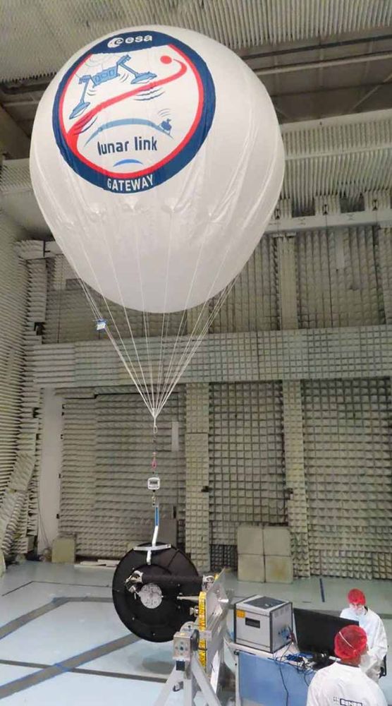 Ballon avec logo "lunar link gateway" de l'ESA, suspendu dans une salle de test, avec des techniciens en blouses blanches et charlottes rouges.