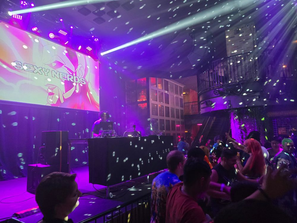 Picture of Sexy Nerds rave event, showing the stage with a projector screen with SEXY NERDS and an anime scene on it. A DJ is playing music to a dancing crowd with disco ball lighting. 