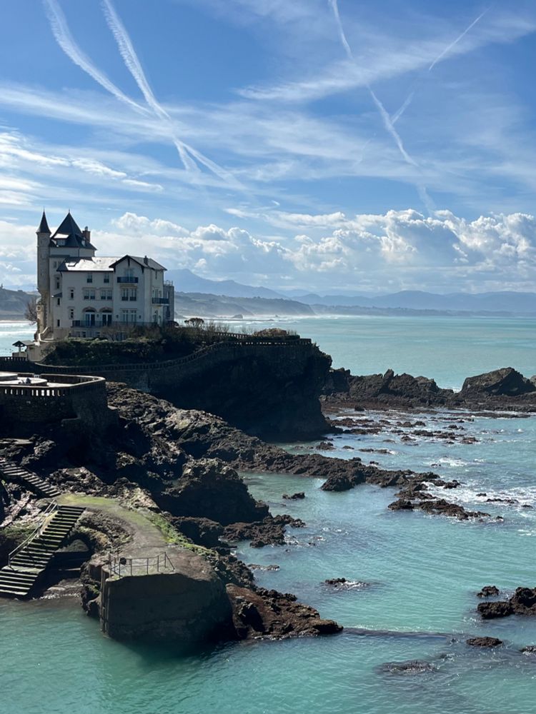 Villa Belza is a white building perched on the rocky coast of Biarritz. The Pyrenees mountains are visible in the far distance. 