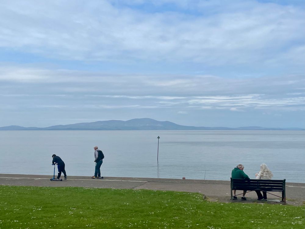 Two people are using scooters and two people sit on bench in Silloth, Cumbria. The hills of Dumfries and Galloway are outlined to the rear of the calm waters of the Solway Firth. A pole stands in the water in the centre of the photo. 