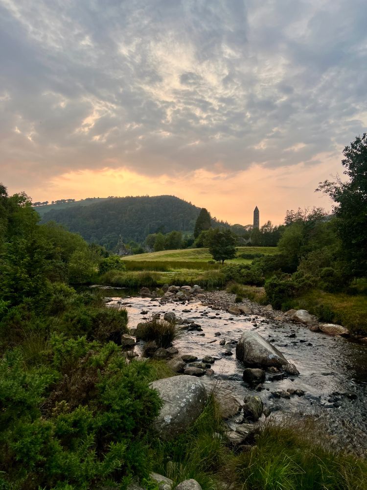 The golden-pink sun sets behind a rolling green hill in the background. Greenery frames the foreground, through which the Glendasan River runs freely over boulders large and small. 