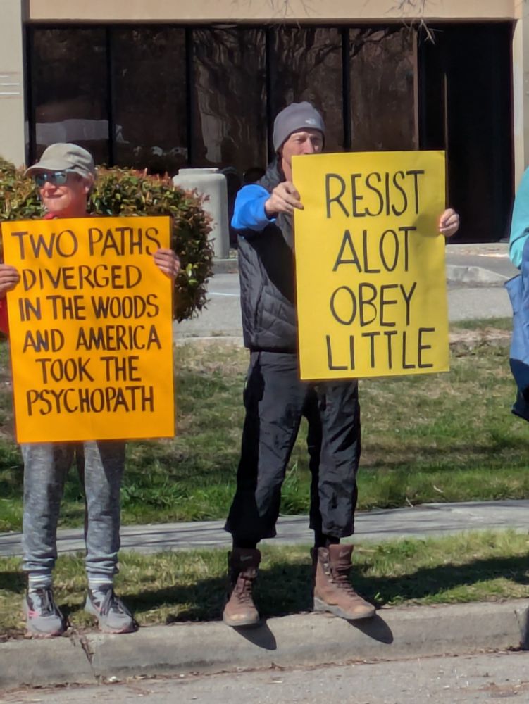 Protesters holding sign says: Two paths diverged in the woods and America took the psychopath; other sign says Resist a lot, obey little, outside Tesla showroom Santa Rosa, 