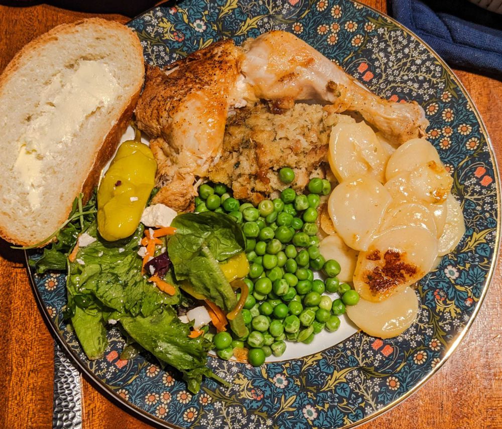 A dinner plate with roasted chicken, stuffing, potatoes, peas, salad, and bread with butter.