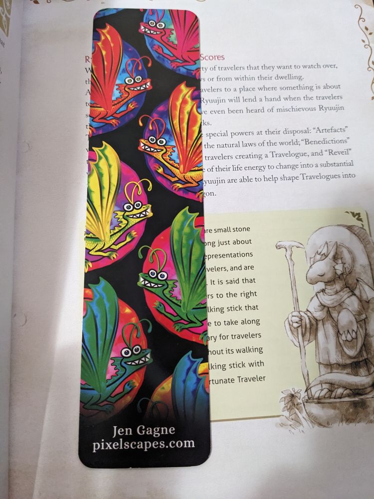 A bookmark of very colourful slightly derpy dragons with Jen Gagne, pixelscapes.com at the bottom. It rests on a page of the Ryuutama rulebook.