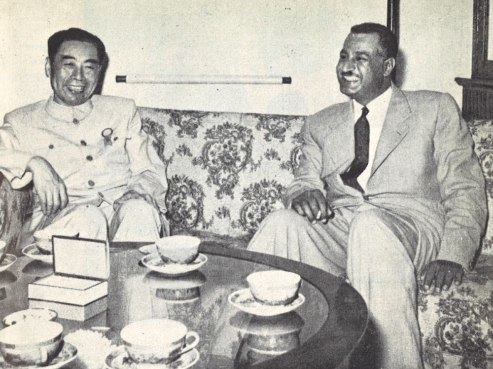 Zhou Enlai (izquierda) charla junto a Gamal Abdel Nasser (derecha). Los dos parecen muy divertidos por algo que se está diciendo durante la conversación. Ante ellos hay una mesita de café con varias tazas y una caja cuya tapa está abierta.