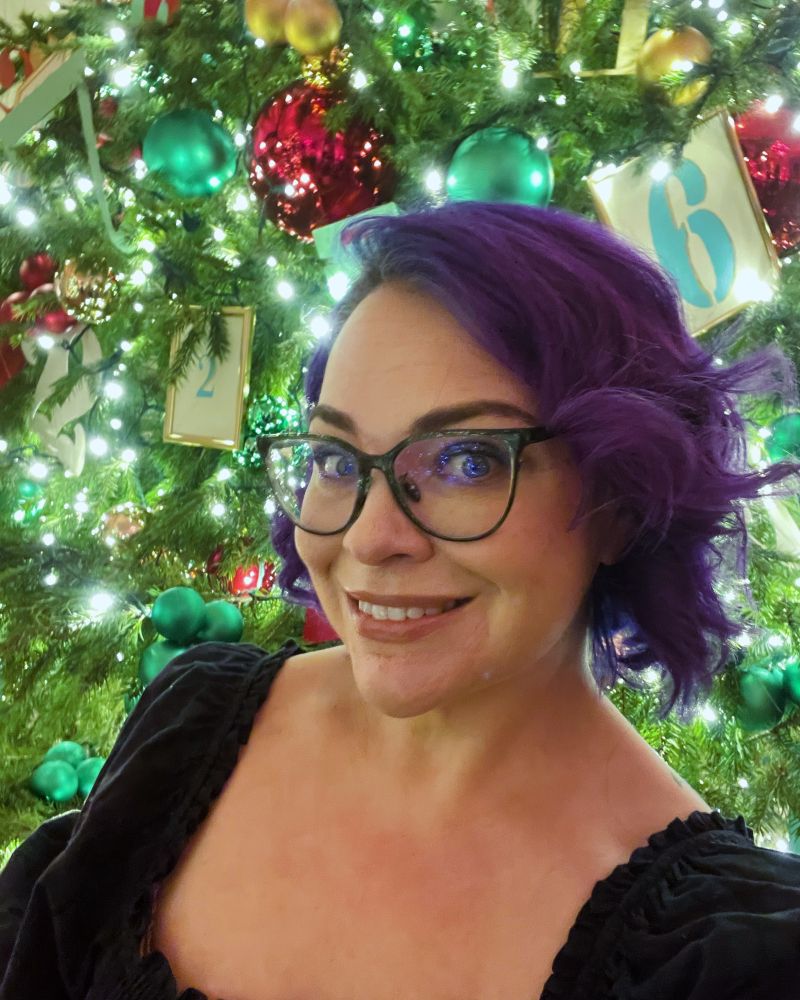Selfie of white-presenting wan wearing glasses with styled, chin-length purple hair in front of a lit Christmas tree.