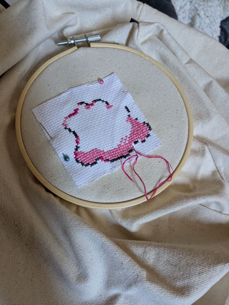 A photograph of a cross stitch work in progress showing outline stitches in black (DMC 360) and blocks in pink (DMC 3806, 3804). There is a square of white Aida on a cream canvas bag, the cross stitch circled with a light wooden frame.
