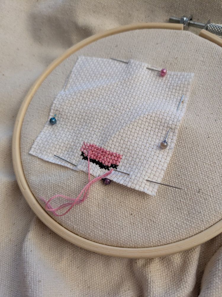 A photograph of a cross stitch in progress. Pictured is a undyed canvas bag with an embroidery hoop sectioning off a piece, a small piece of Aida pinned to the canvas. There is a small patch of stitches completed in pink, the bulk of the stitches, and black, lining the bottom