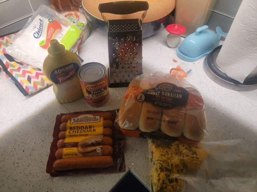 Yellow mustard, a can of chili, hot dog buns, Johnsonville Cheddar brats, grated cheese, and a cheese grater