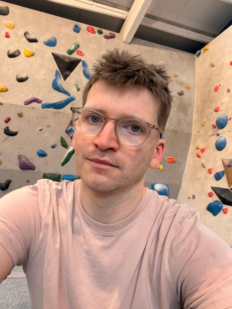 Selfie, I’m wearing a pale pink top, my hair is freshly cut short. Background of a bouldering gym