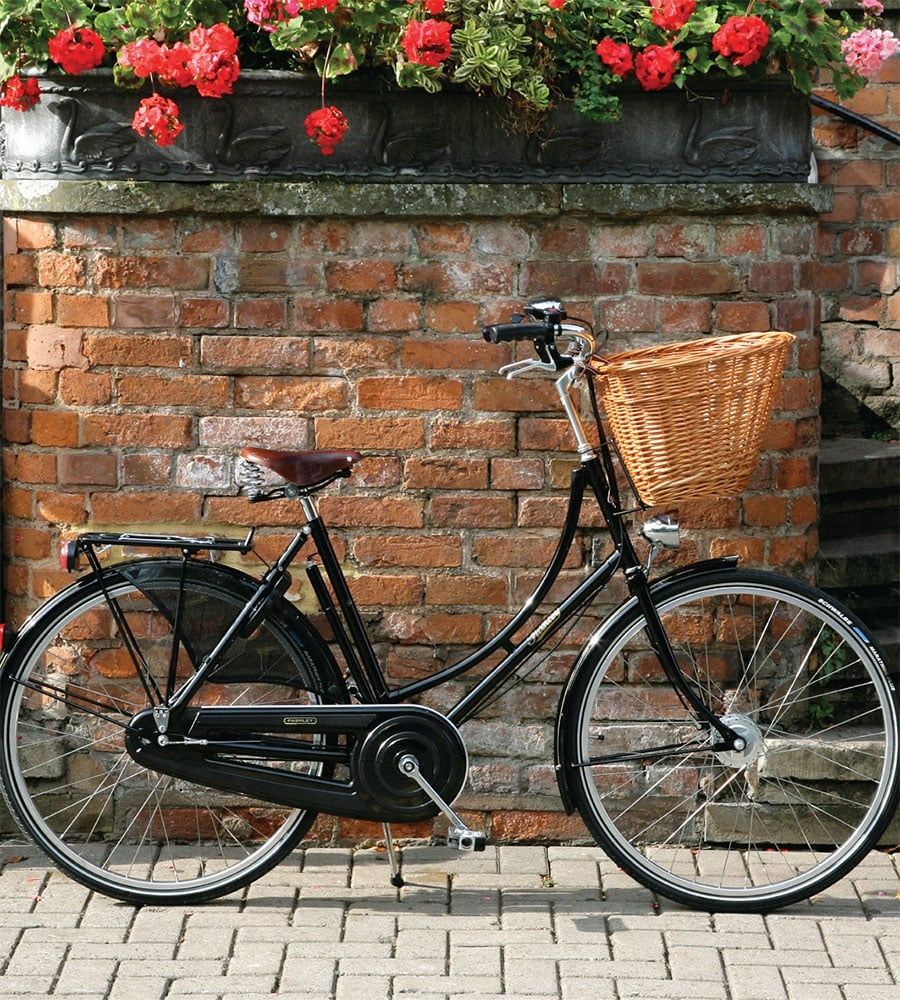 Photo of a Pashley brand Princess bicycle that looks like it hasn't been ridden so it is new and glossy. It's a Dutch style bike that you sit upright on like a horse, with a whicker basket up front and a pannier rack at the back plus a sprung leather seat. It looks like it is from the 1940's, and is rather lovely. 