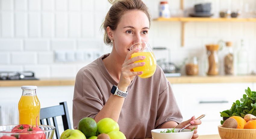 Person sitting at a kitchen table enjoying a glass of orange juice with fresh fruits and a bowl of salad nearby.
