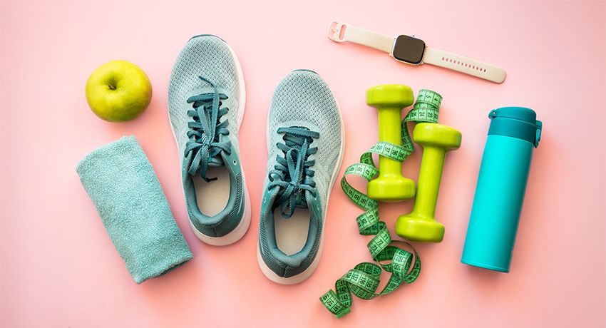 Pair of light blue running shoes with teal laces next to a green apple and rolled towel on a pink background.