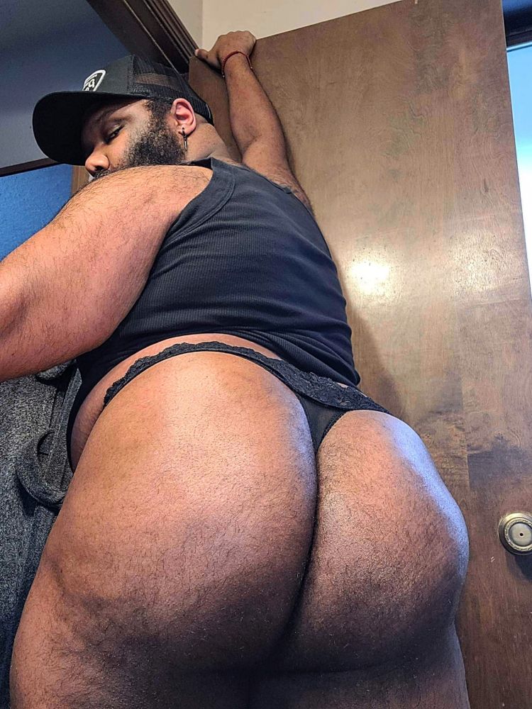 A bearded trashpanda stands facing away in an open doorway. Clad only in a trucker hat, a black tank, & a black lace thong almost completely swallowed up by his insanely fat hairy ass cheeks, his smoldering gaze pins you as he looks at you over his shoulder.