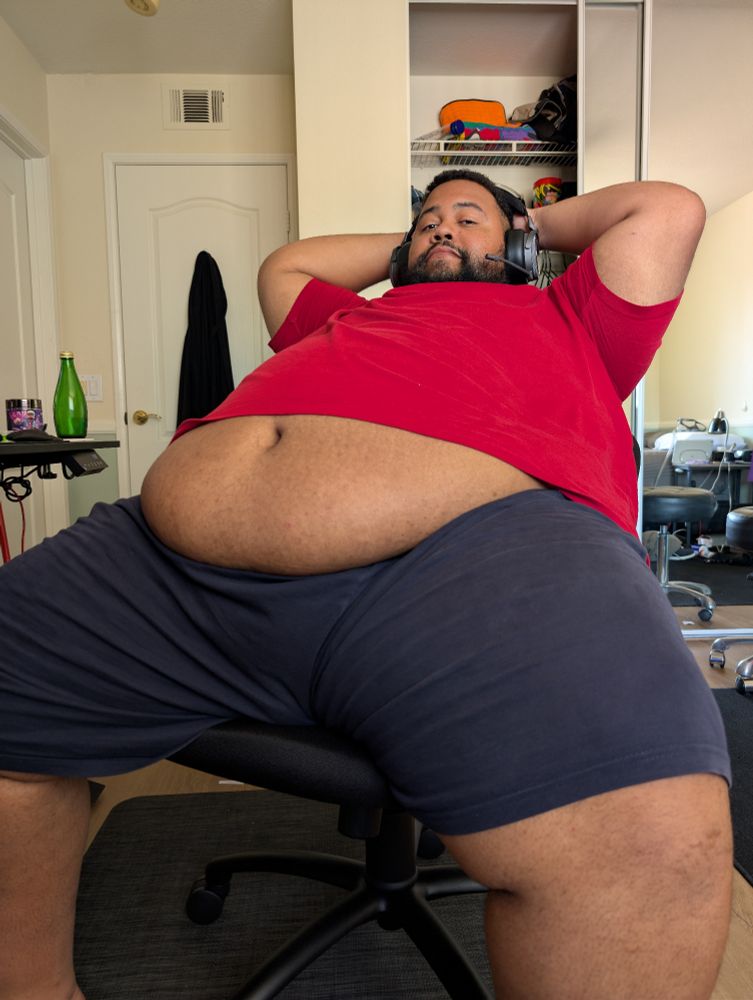 Superchub wearing a red shirt and blue shorts sitting in an office chair leaning back with his hands behind his head, belly exposed showing off his navel and softness 