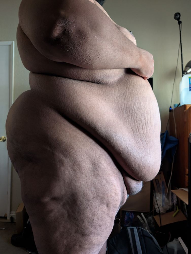 Side profile of a superchub standing nude showing off his rolls and folds 