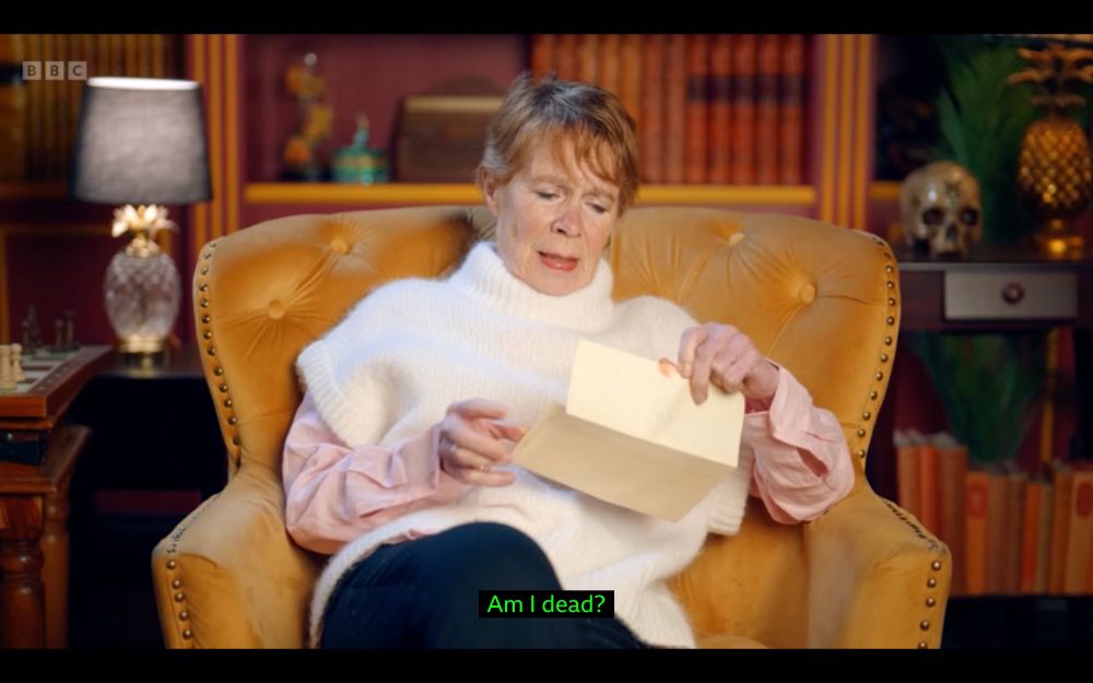 Celia Imrie on Celebrity Traitors opening a sealed letter and reading it. The subtitle on screen says "Am I dead?"