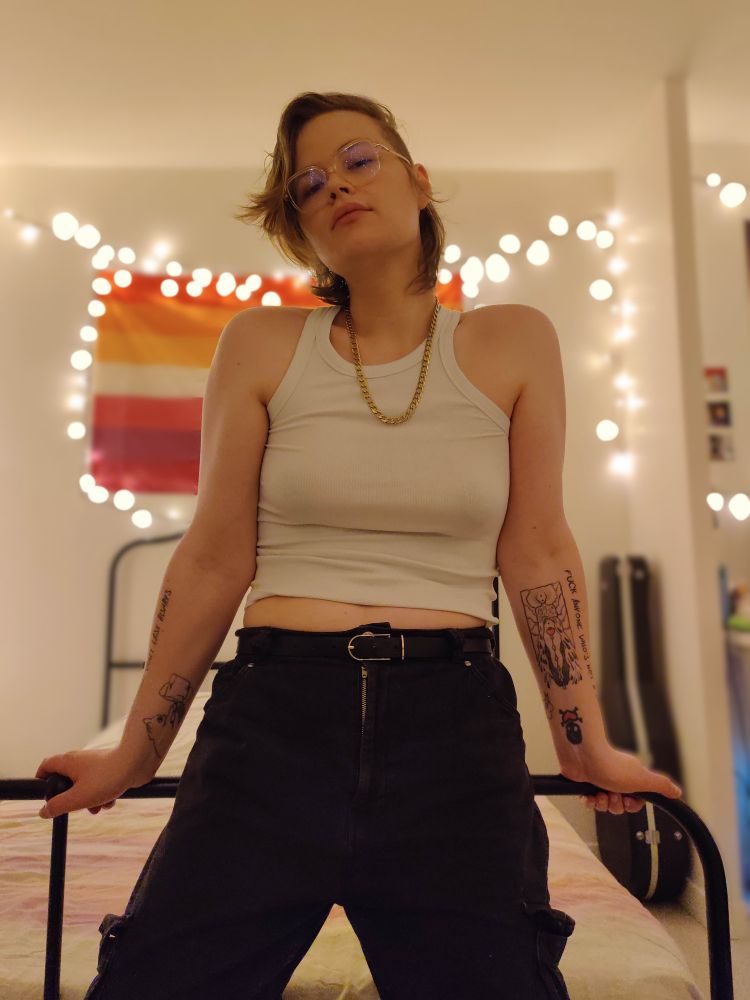 photo autoportrait dans ma chambre, je porte un débardeur côtelé blanc de gouine, un pantalon noir avec une ceinture, ma chaîne dorée, mes lunettes roses transparentes et je sais pas pour qui je me prends mais je regarde la caméra en mode séducteurice ptdr. je flex mes bras de manière casual (mais un peu cramée) pour les lesbiennes qui tomberont sur cette photo. je suis debout/assise contre le cadre de mon lit. en arrière plan : ma guitare dans sa caisse protectrice près de mon lit, un drapeau lesbien accroché au mur ainsi qu'une guirlande lumineuse encadrant le drapeau avec un effet de bokeh