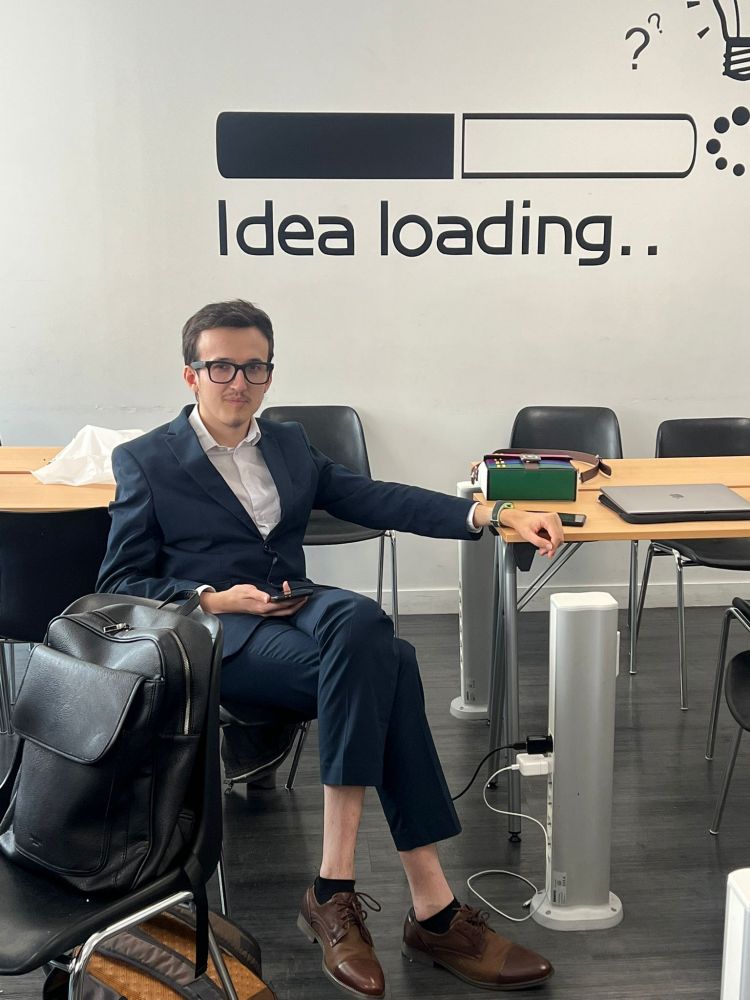 Photo de pessi distingué posé à l'aise dans une classe, la main gauche sur la table à côté de lui et les jambes croisées en costard.