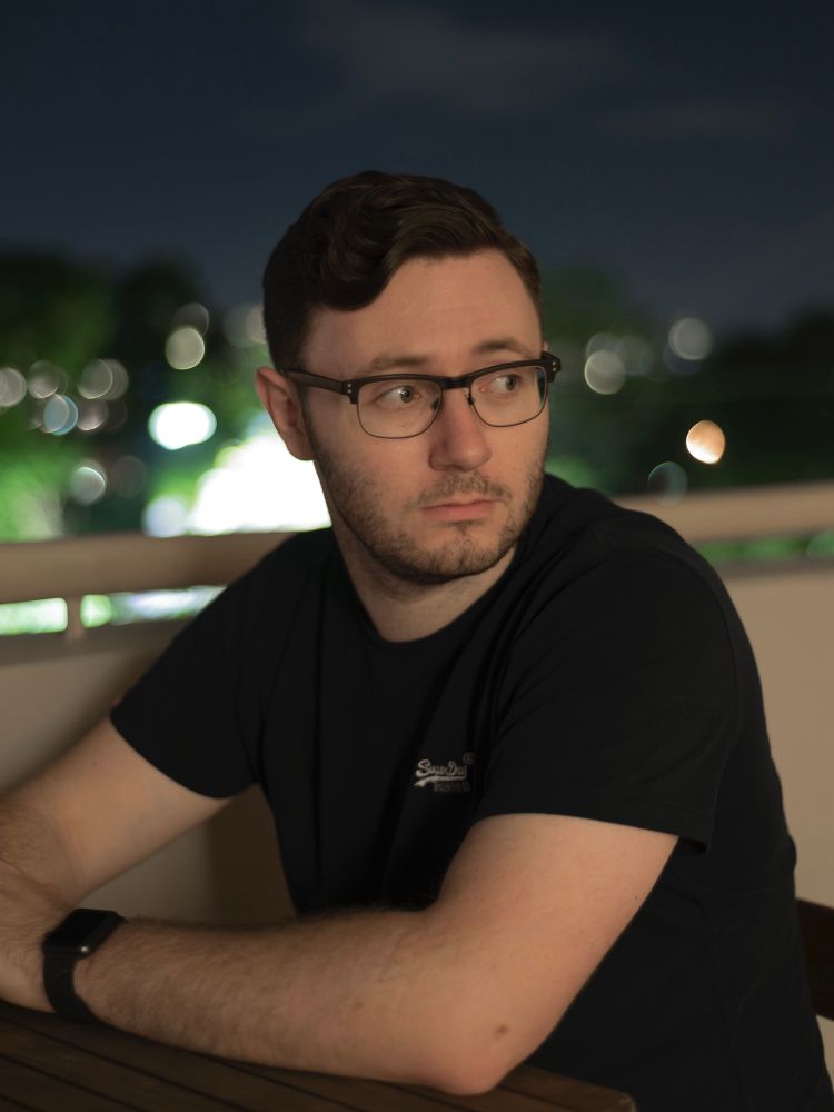 Photo of me wearing a navy blue t-shirt looking to my left with an out of focus night city background.