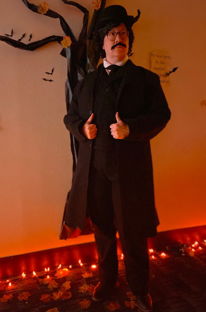 A full body shot of Sage in a black vest, pants, white dress shirt, cravat, jacket, black curly wig, mustache, and derby. In the background is orange light and a spooky paper tree