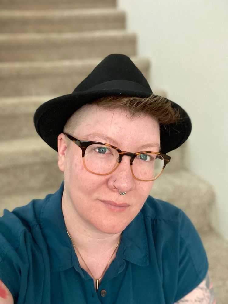Sage wears a black felt fedora, thick rimmed glasses, and a teal short-sleeved button-up shirt