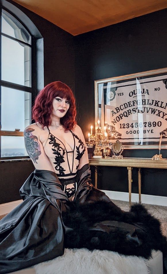 photo of a woman sitting on a white fur rug in a luxurious black room with gold ceiling. there is a large window behind her and on the other side a gold table with candleabra and large ouija board on the wall while she sits with a slight smirk and wears a nude coloured bodysuit with black flowers embroidered on it 