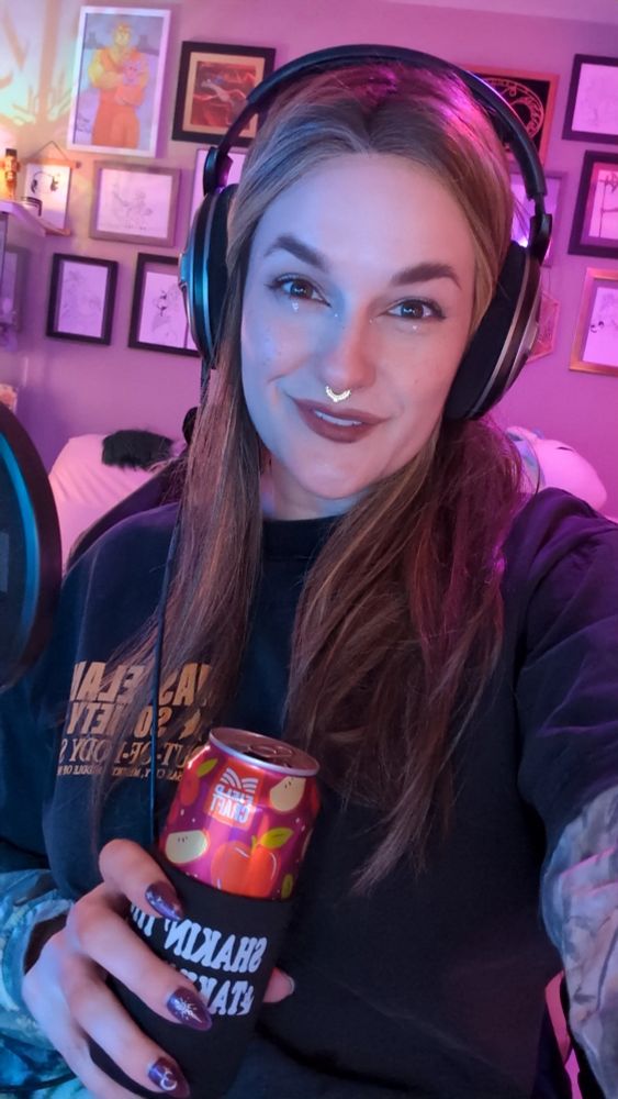 Selfie of Nika holding a cider can and wearing headphones while smiling at the camera in a bisexually lit room