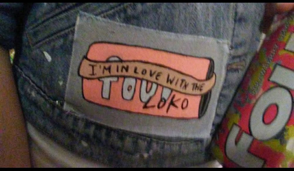 A patch safety-pinned onto a jacket depicting a can of Four Loko with the text "I'm in love with the Loko" written on it. There is also a can of Four Loko being held up right next to the patch.