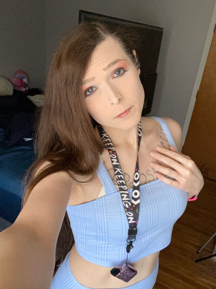 Emily Love stands with a bed and tv behind her. She’s wearing a plaid blue crop top and a blue plaid skirt.