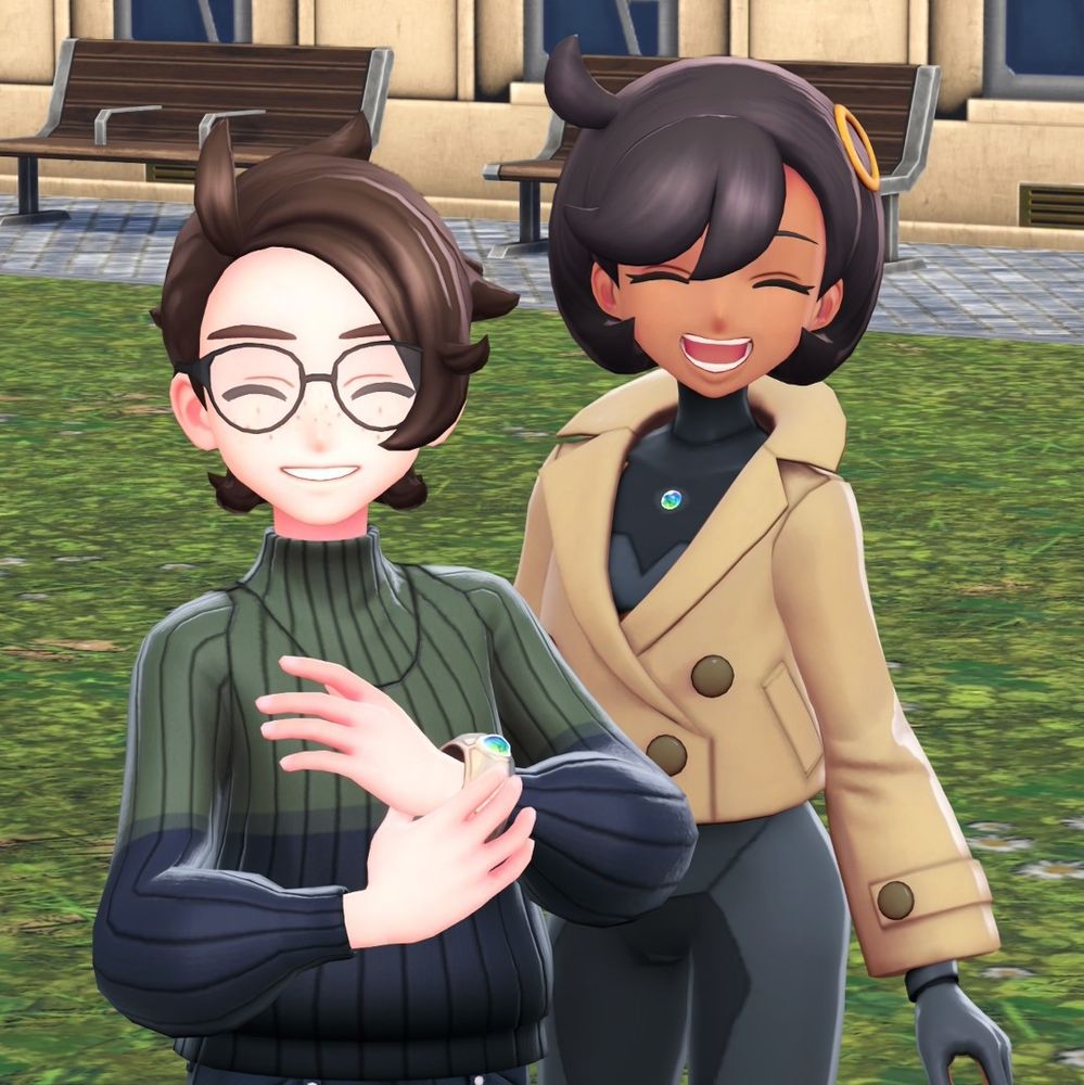 Videogame screenshot of a selfie with a man and woman in a park. The man has brown hair, freckles and glasses, wearing a green/blue jumper and smiling. The woman has short black hair and wearing a black bodysuit and tan trench coat, also smiling.