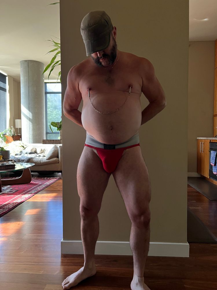 looking good in a red jock, nipple clamps, and a downturned hat in a lit living room 
