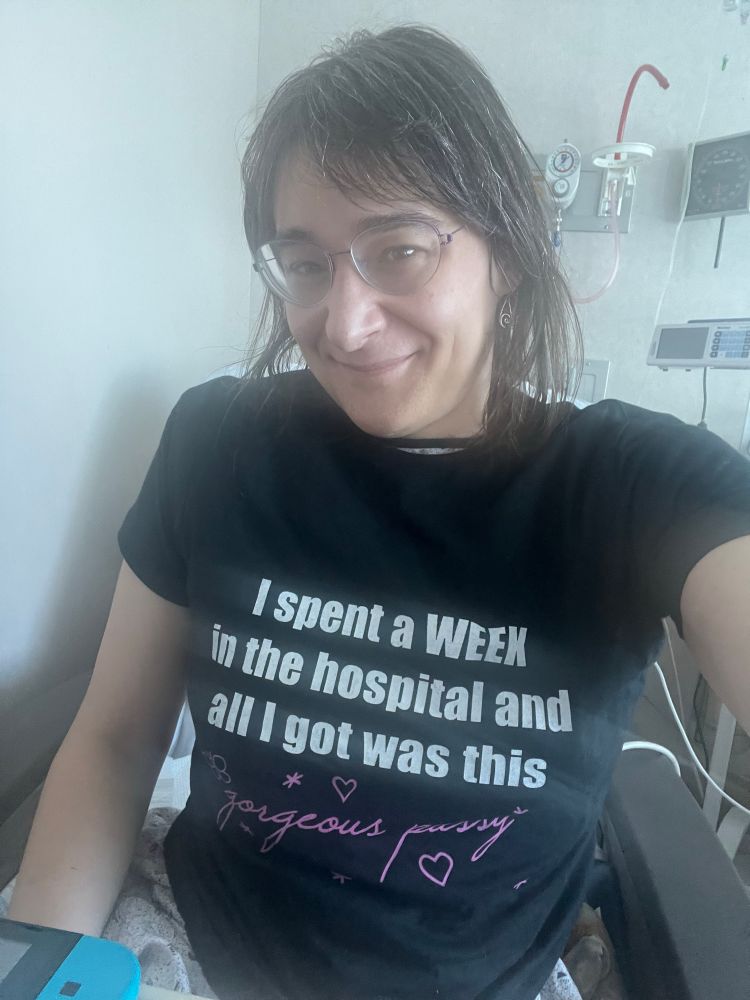 static in a tshirt that is too big for her. it's black and says, in impact font, "I spent a WEEK in the hospital and all I got was this" followed by, in pink script with little stars and hearts surrounding it, "gorgeous pussy"