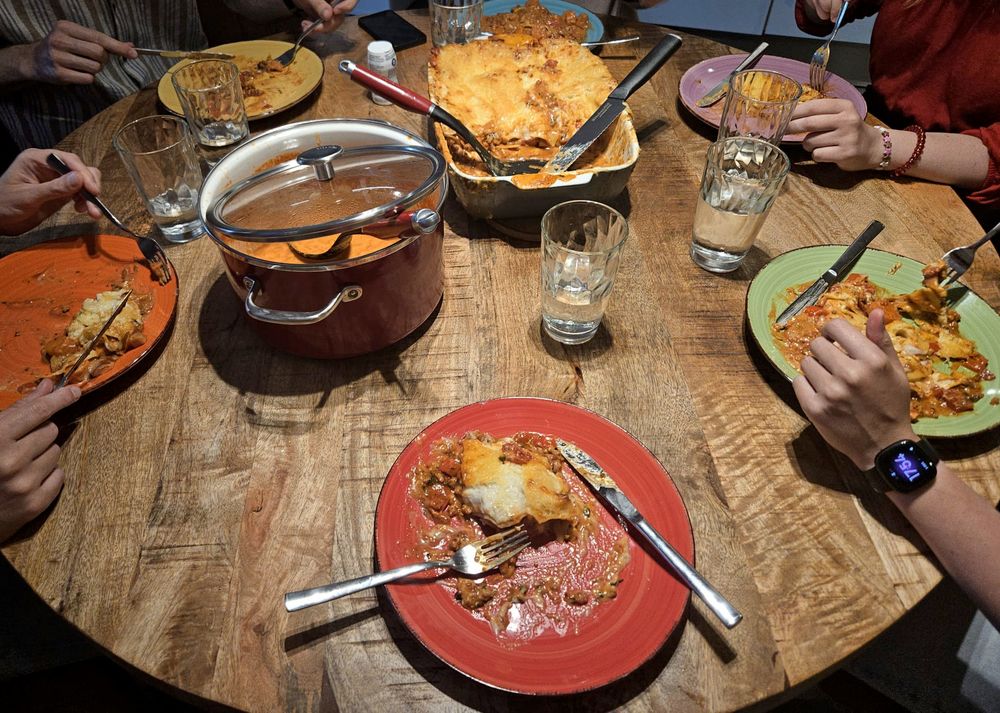 A round dinner table with six plates in the colors of the rainbow with lasagne on them.