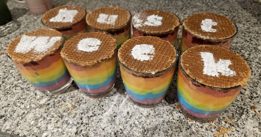 Eight glasses filled with six layers of yoghurt in the colors of the rainbow, topped off with a stroopwafel. On top, letters made out of powdered sugar spell out RustWeek.