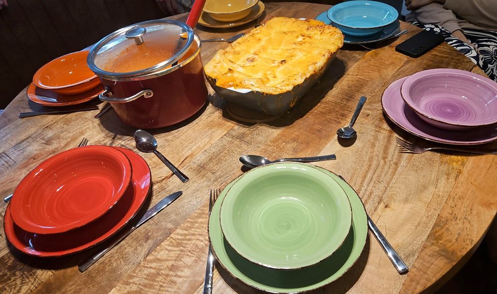 A round dinner table with six matching plates and bowls, each in a different color of the rainbow. In the middle is a pot of (bell pepper) soup and a big lasagne dish.