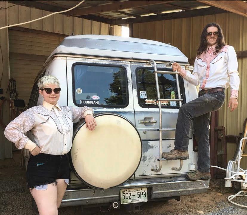 Two people posing at the back of a battered gray and white camper van with stickers on the windows. On the left, a woman with short hair leans against the spare tire with one hand on her hip. She has pale skin & short bleach-blonde hair and is wearing sunglasses. short black shorts, and an offwhite cowboy shirt with floral accents. On the right, hanging from the ladder on the back of the van, is a man with pale skin, a beard, and long, curly brown hair. He is wearing sunglasses, jeans, and a matching cowboy shirt with pink floral accents. 