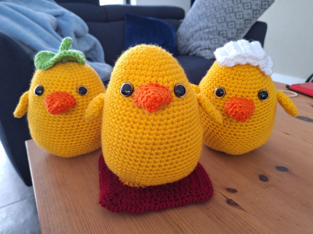 Three ovoid yellow crochet ducks with orange beaks. One has a green leaf on its head, one sits on a red cushion, and one has a folded white towel on its head. Adapted from pattern by KnitKnot2Know.