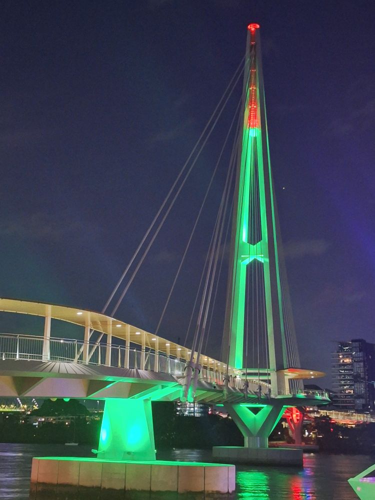 A pedestrian footbridge in front of the Brisbane skyline at night, suspended from a huge suspension tower and lit up in green