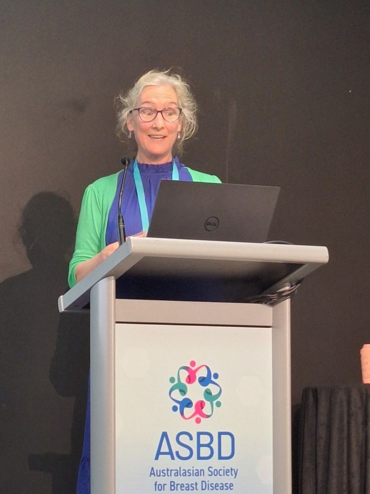 Dr Virginia Baird with an animated expression standing at a podium bearing the Australasian Society of Breast Disease logo.