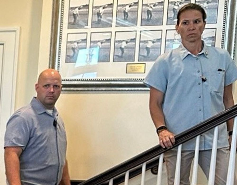 closeup of a portion of the OP image showing two people in light blue shirts ascending or paused on a staircase the person above is directly facing the camera with a serious, concerned face. the person below is turned 3/4 toward the camera and also looking at it with a serious, concerned face