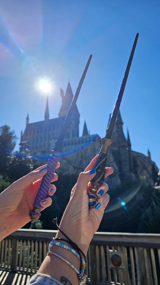 Image shows myself and my sister holding up 2 wands from Hogwarts Legacy with Hogwarts castle in the background at Universal studios Orlando. 