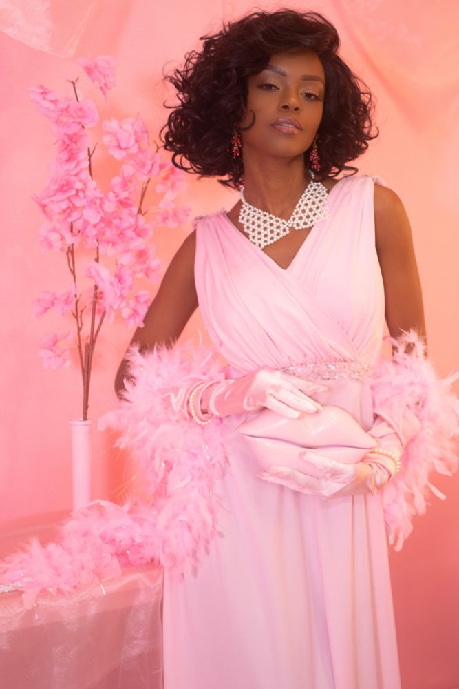 A striking Black woman exudes vintage glamour in a soft pink gown with pearl embellishments, satin gloves, and a feathered boa. Her sultry gaze and confident posture complement the luxe details—crimson earrings, a pearl statement necklace, and a glossy, lip-shaped clutch. Set against a dreamy pink backdrop with cherry blossoms, the scene radiates Old Hollywood elegance with a modern, seductive twist.