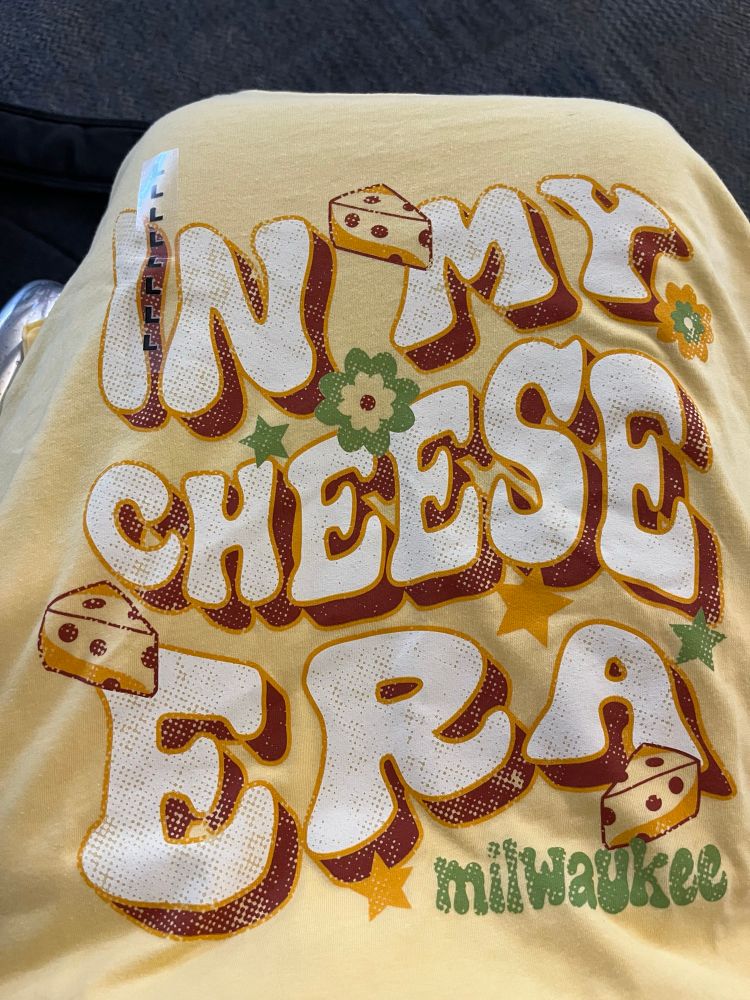 Close up of a yellow tee shirt with the phrase “in my cheese era Milwaukee” in funky, 70’s style lettering in brown, white, and green, with flower and star accents and wedges of Swiss cheese. Illustrates my love of both the dairy product and Taylor.