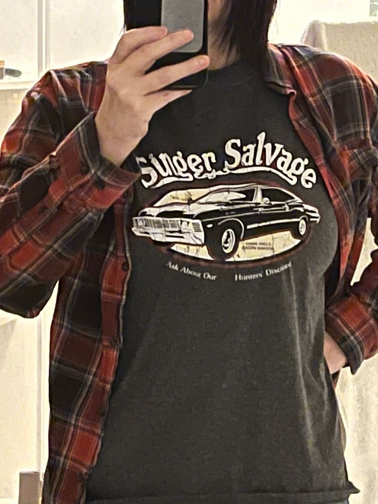 Grumpy old woman wearing a flannel shirt with a gray t-shirt underneath that reads “Singer Salvage/ask about our hunter’s discount” with an image of a black 1967 Impala, the signature car from the TV series Supernatural, ya idjit.
