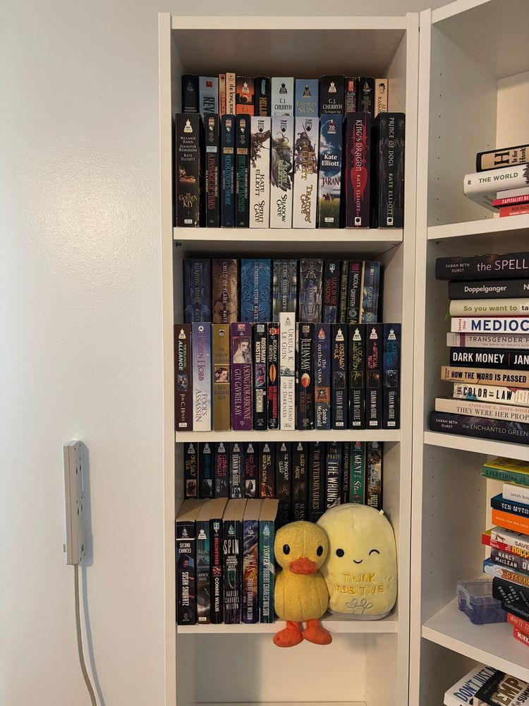 A bookshelf; the top three shelves are packed with science fiction and fantasy mass market paperbacks, double stacked using shelf risers. On the third shelf down, there are two stuffies: one duck with its feet dangling and a winking light bulb that says “think positive”.